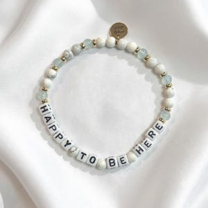 May include: A beaded bracelet featuring white marbled beads, translucent light blue beads, and small gold-toned spacers. The bracelet includes white square letter beads spelling out the phrase HAPPY TO BE HERE and a small round gold-toned metal charm.