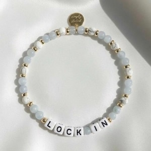 May include: A beaded bracelet featuring light blue and white beads, interspersed with gold-colored accents. The bracelet includes white letter beads spelling "LOCK IN" and a small gold charm with the text "simply stated".