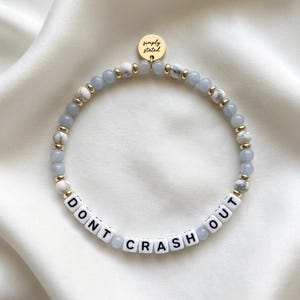 May include: A beaded bracelet featuring light blue and white marbled beads, small gold-toned spacers, and white letter beads spelling DONT CRASH OUT. A round gold-toned charm at the top is engraved with the text Simply Stated.
