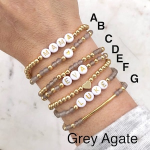 May include: Seven bracelets stacked on a wrist, each with a different name spelled out in gold letters on white beads. The bracelets are made with grey agate beads and gold beads. The names are "MAMA", "EVA", "LUKE", and a heart charm. The bracelets are labeled A through G.