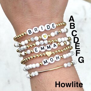 May include: Seven stretch bracelets labeled A through G, featuring white howlite beads, gold-tone metallic beads, and white letter beads spelling BRIDE, EMMA, and MOH. Some bracelets include gold-tone heart charms or a gold-tone bar accent piece.