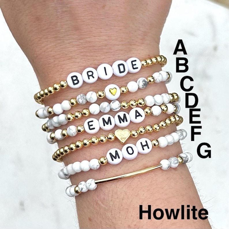 May include: Seven stretch bracelets labeled A through G, featuring white howlite beads, gold-tone metallic beads, and white letter beads spelling BRIDE, EMMA, and MOH. Some bracelets include gold-tone heart charms or a gold-tone bar accent piece.
