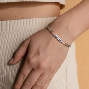 Puede incluir: Una pulsera de cuentas con la palabra "MAMA" escrita con cuentas de letras blancas. La pulsera presenta cuentas grises y rosas, y detalles dorados. La pulsera se lleva en la muñeca.
