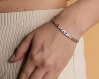 May include: A beaded bracelet with the word "MAMA" spelled out in white letter beads. The bracelet features gray and pink beads, and gold-colored accents. The bracelet is worn on a wrist.