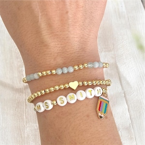 May include: Three beaded bracelets stacked on a wrist. One bracelet has light blue beads, another has a gold heart charm, and the third spells "MS SMITH" with a pencil charm. The bracelets are gold in color.