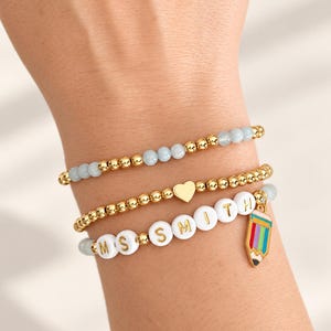 May include: A set of three beaded bracelets. One features light blue and gold beads, another has gold beads with a central gold heart charm, and the third has white letter beads spelling MS SMITH with a colorful pencil charm attached to the end.