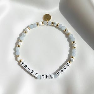May include: A beaded bracelet with light blue, white, and gold accents. The bracelet features the words "TRUST THE PACE" spelled out in white letter beads. A small gold charm is attached.