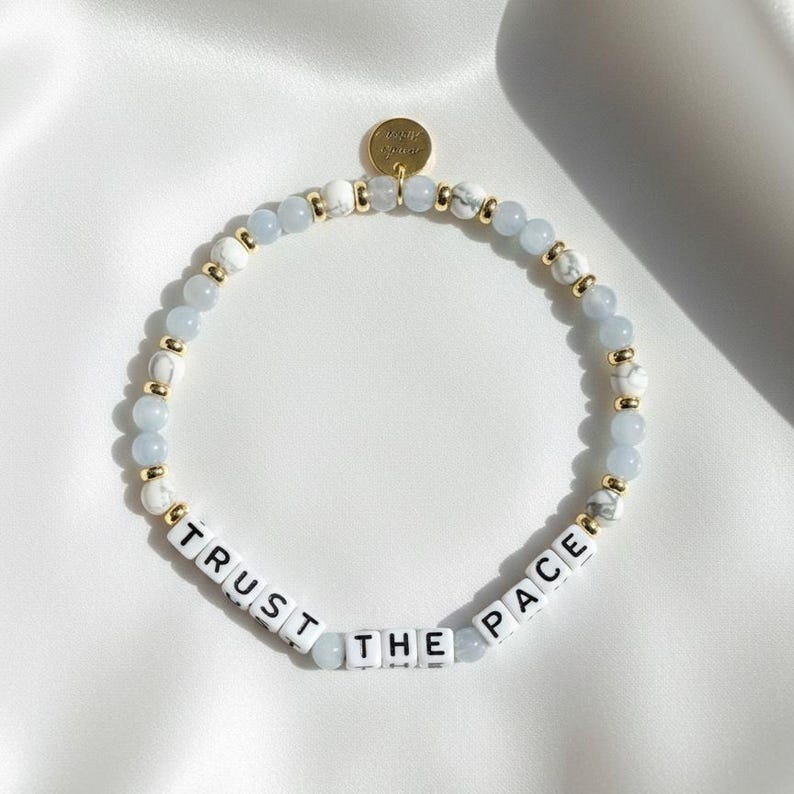 May include: A beaded bracelet with light blue, white, and gold accents. The bracelet features the words "TRUST THE PACE" spelled out in white letter beads. A small gold charm is attached.