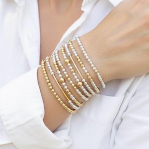 Puede incluir: Una colección de pulseras de cuentas doradas y plateadas adornan una muñeca. Las pulseras varían en diseño, algunas con barras de oro macizo y otras que consisten enteramente en pequeñas cuentas redondas. La joyería se muestra contra una camisa blanca.