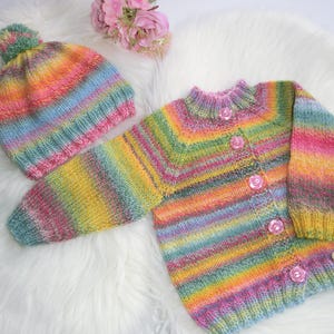 Könnte beinhalten: Eine regenbogen gestreifte Strickjacke für Babys mit passender Mütze. Die Strickjacke hat einen Knopfverschluss und die Mütze hat einen Bommel oben.