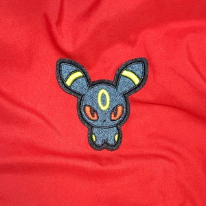 May include: An embroidered patch of a Pokemon character, Umbreon, in dark blue with yellow accents, on a red fabric background.