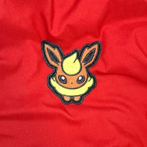 May include: A red fabric background with a black and yellow embroidered patch of a cartoon character. The character has brown fur, yellow hair, and black eyes.