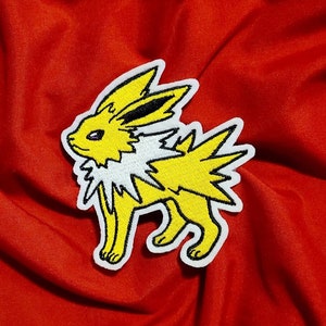 May include: Yellow and white embroidered patch of a Pokemon character,  Eevee's evolution, Jolteon. The patch is on a red fabric background.