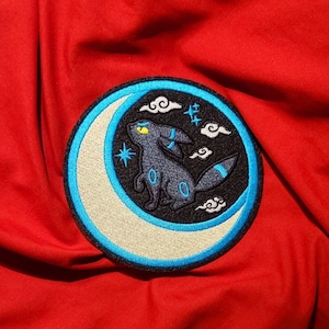 May include: A blue and white embroidered patch featuring a black and blue Pokemon character standing on a crescent moon with clouds and stars in the background.