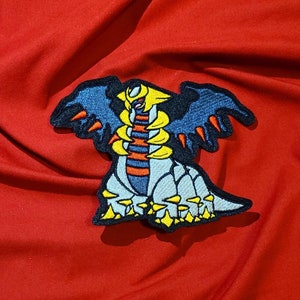 May include: Embroidered patch depicting a stylized creature with a yellow head, blue wings, and a grey body. The patch is set against a red background. The design includes orange and yellow accents, creating a vibrant and detailed image.