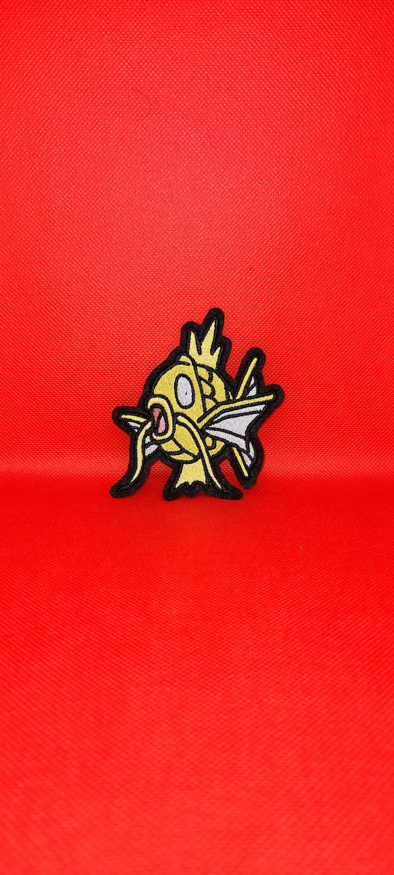 May include: A yellow and black embroidered patch of a cartoon fish with a crown on its head. The fish has a large mouth and is swimming to the right.