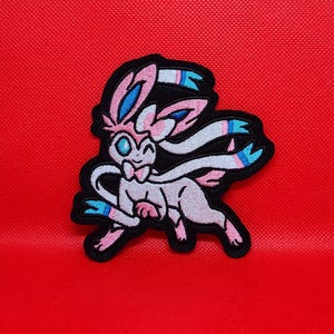 May include: Pink and blue embroidered patch of a Pokemon character, a mythical creature with long ears and a tail, with a black outline.