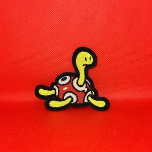May include: A yellow and red embroidered patch of a cartoon turtle with a shell and a long neck.
