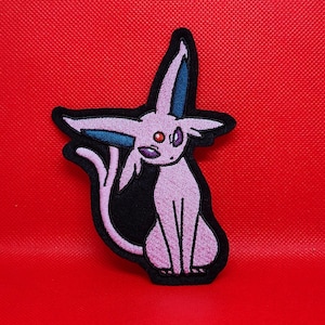 May include: A pink and blue embroidered patch of a Pokemon character, Espeon, sitting on a black background.