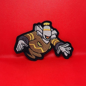 May include: A brown and yellow embroidered patch of a cartoon robot with a single eye and a halo. The robot has a striped body and is in a flying pose with its arms outstretched.