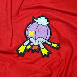 May include: A colorful embroidered patch featuring a cartoon-style character with a purple body, yellow cross-shaped eyes, and yellow feet. The patch is set against a vibrant red fabric background.