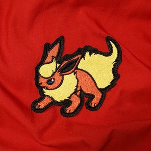 May include: Embroidered patch of a yellow and orange Pokemon character with black outlines. The character has a fluffy tail and is standing on all fours.