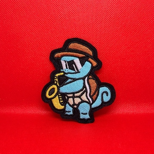 May include: A blue cartoon turtle wearing a brown hat and sunglasses plays a yellow saxophone. The turtle is embroidered on a black fabric patch.