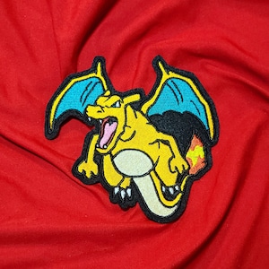 May include: A yellow and orange embroidered patch of a cartoon dragon with blue wings. The dragon is facing forward with its mouth open and has a flame coming out of its tail.