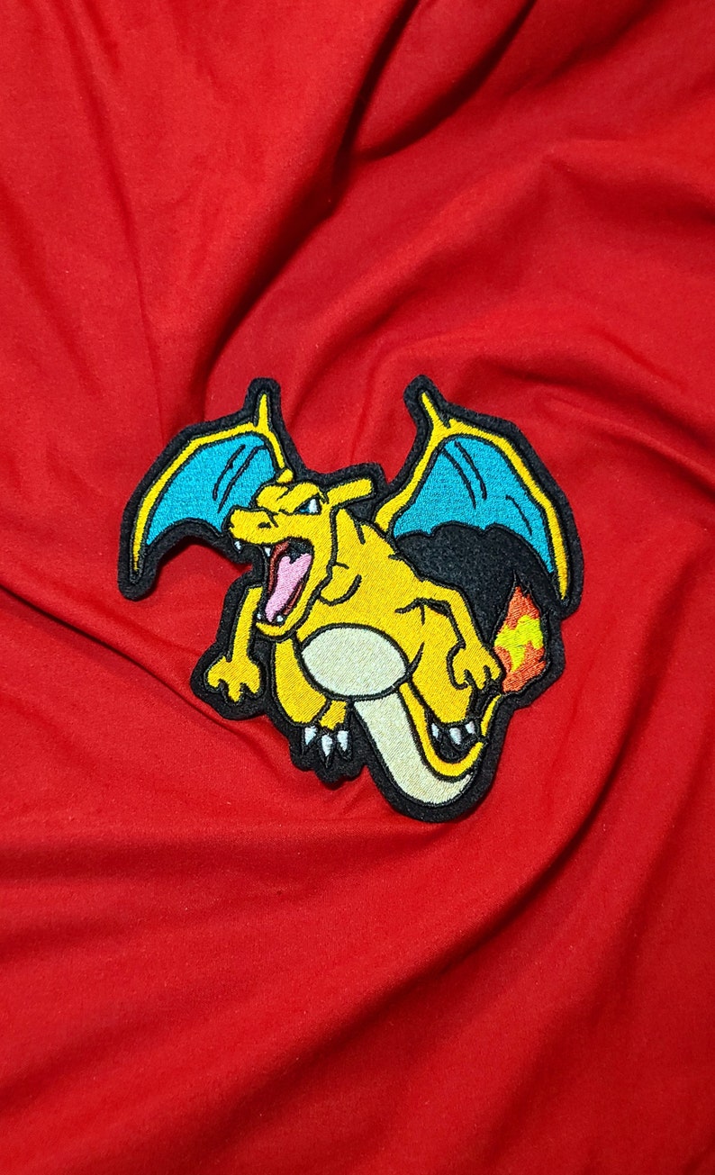 May include: A yellow and orange embroidered patch of a cartoon dragon with blue wings. The dragon is facing forward with its mouth open and has a flame coming out of its tail.