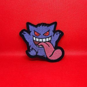 Peut inclure: Écusson brodé représentant un fantôme de dessin animé violet avec un large sourire, des yeux rouges et une longue langue rose. L'écusson est sur un fond rouge vif. Idéal pour les vêtements et accessoires.