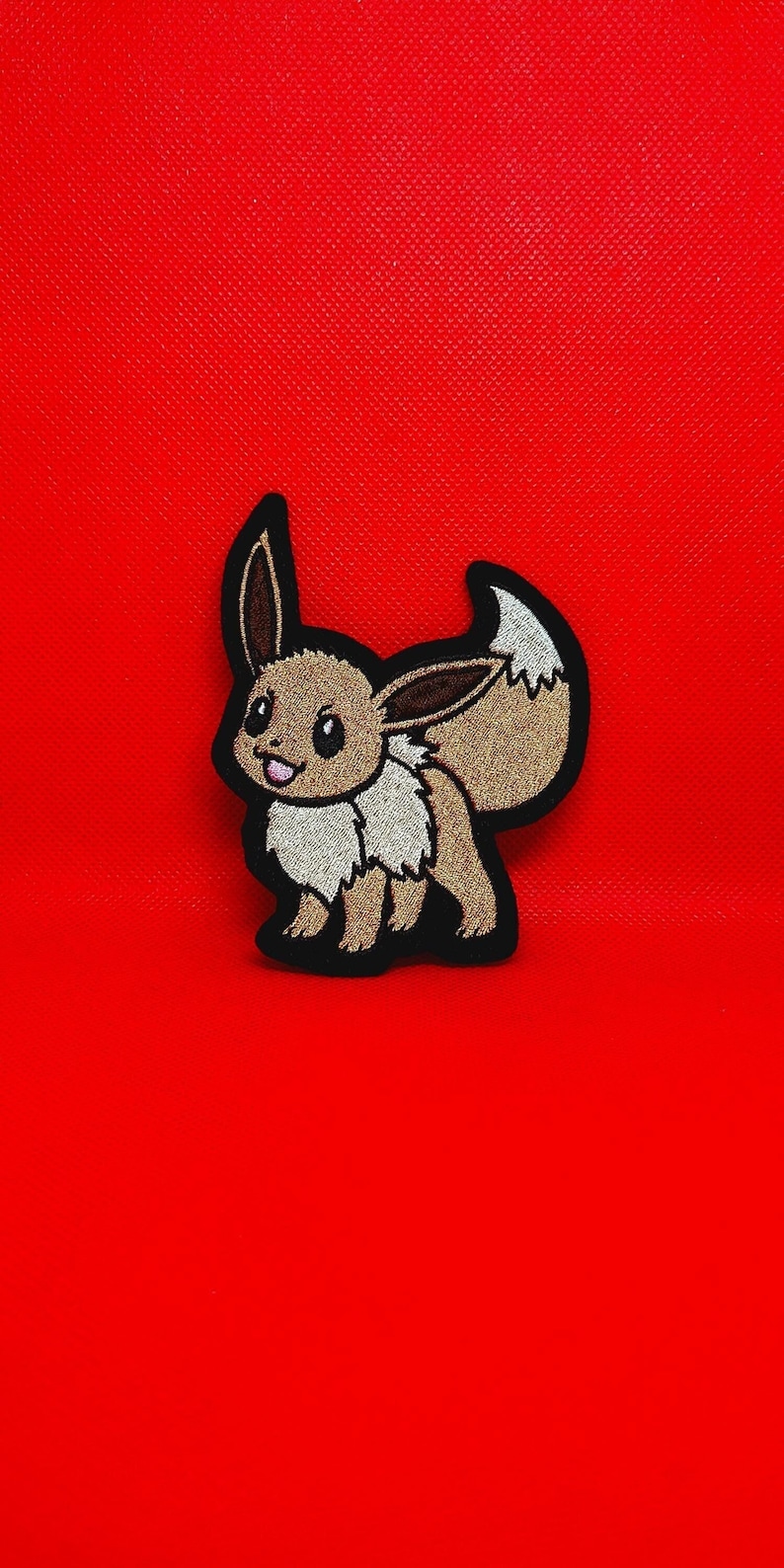 Pu&ograve; includere: Toppa ricamata di un personaggio Pok&eacute;mon Eevee marrone con contorno nero. L'Eevee &egrave; in piedi su uno sfondo rosso.