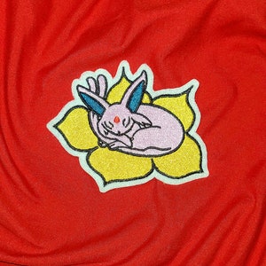 May include: An embroidered patch featuring a sleeping Eevee Pokemon in pink and blue, resting on a yellow flower.