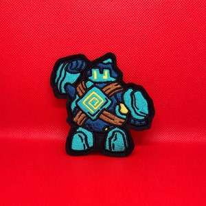 May include: An embroidered patch featuring a robot character in blue and brown, with a yellow square on its chest.