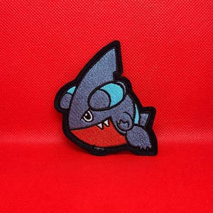 May include: An embroidered patch featuring a stylized dark blue creature with a prominent dorsal fin, light blue accents, and a bright red lower section. The character has a small white tooth visible and is outlined in thick black thread.