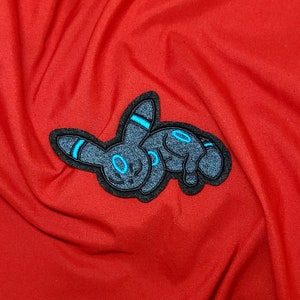 May include: Black and blue embroidered patch of a Pokemon character,  Eevee, on a red fabric background.