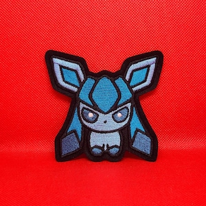 May include: An embroidered patch featuring a stylized creature with large pointed ears, a light blue face, and dark blue accents. The design is outlined in thick black thread, showcasing a symmetrical character with wide eyes and a small nose.