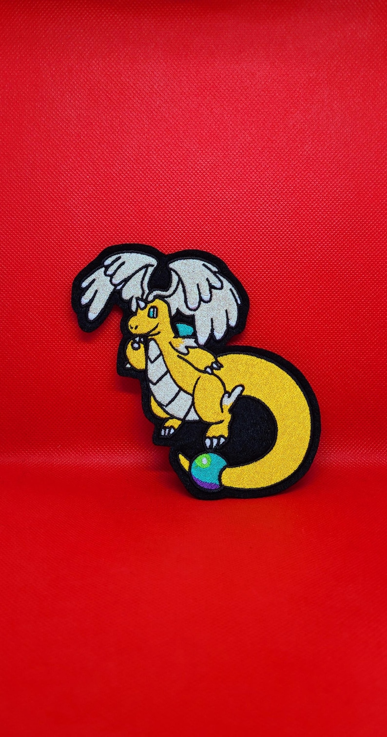 Peut inclure: &Eacute;cusson de dragon jaune avec des ailes blanches et un contour noir, sur fond rouge. Le dragon a une queue incurv&eacute;e et un orbe multicolore &agrave; l'extr&eacute;mit&eacute;. L'&eacute;cusson est probablement destin&eacute; aux v&ecirc;tements ou aux accessoires.