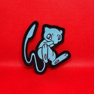 May include: A blue and black embroidered iron-on patch featuring a cartoon character with a curled tail. The character is light blue with black outlines and white eyes.