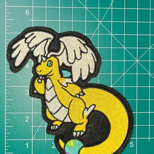 Peut inclure: &Eacute;cusson brod&eacute; repr&eacute;sentant un dragon jaune avec des ailes blanches, un contour noir et un globe multicolore. L'&eacute;cusson mesure environ 10 cm de haut et 13 cm de large.