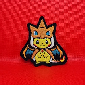 May include: Embroidered patch featuring a yellow Pikachu wearing a Charizard costume. The patch has a black border and is set against a red background. The design includes orange, blue, and white details.