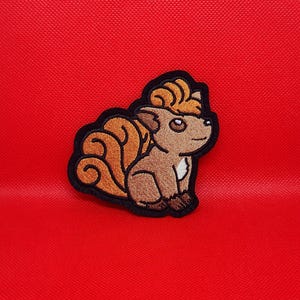Peut inclure: Écusson brodé représentant une créature de dessin animé avec une fourrure marron, des détails orange et une bordure noire. La créature a de grands yeux et une queue touffue. L'écusson est posé sur un fond rouge uni.