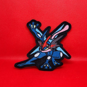 May include: A blue and red embroidered patch of a Pokemon character, Greninja, in a fighting pose. The patch is on a red background.