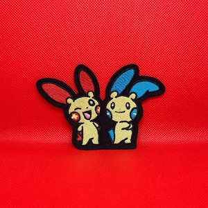 May include: Embroidered patch featuring two cartoon characters with yellow bodies, black outlines, and bunny-like ears. One character has red ears, the other blue. The patch is set against a solid red background.