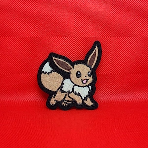 May include: An embroidered patch featuring a brown Eevee character with a cream-colored chest ruff and tail tip. The character has large dark eyes, pointed ears, and a black stitched border outline, presented against a solid red background.