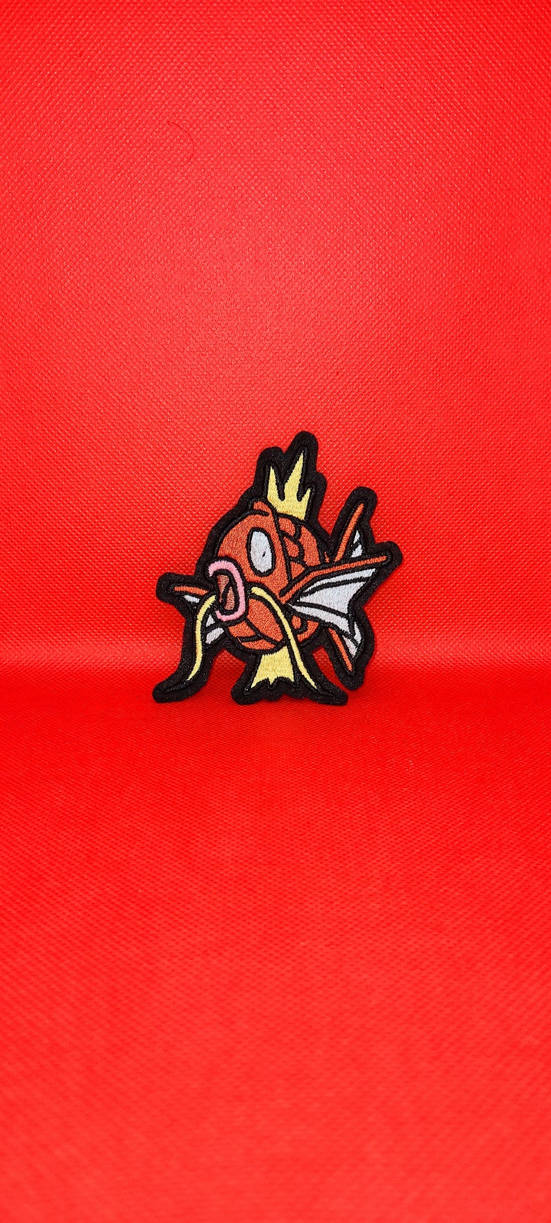 May include: A red and orange embroidered patch of a cartoon fish with a crown on its head. The fish has a yellow mouth and a yellow fin.