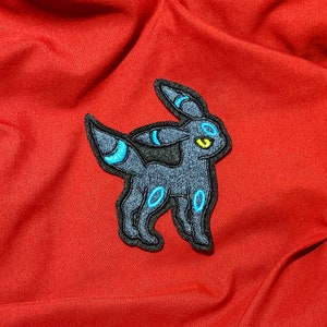May include: An embroidered patch featuring a mythical creature in blue and black, with yellow eyes. The creature is standing on all fours with its tail curled up.