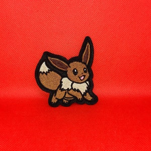 Peut inclure: Un patch brodé représentant un dessin animé d'Eevee, un personnage de Pokemon, en brun et beige avec un contour noir. Le patch est sur un fond rouge.