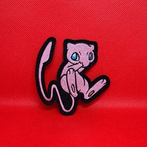 May include: Pink and black embroidered iron-on patch featuring a cartoon image of a pink Pokemon character with blue eyes.