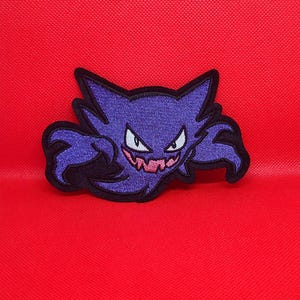 May include: A purple embroidered patch featuring a stylized creature with pointed ears, sharp white eyes, and a wide grin showing jagged pink teeth. The design is outlined in thick black thread, creating a bold, high-contrast appearance.