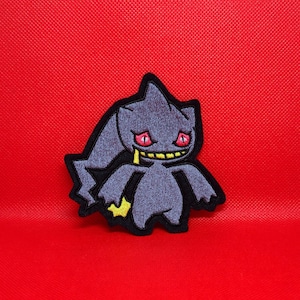 May include: A gray embroidered patch of a cartoon ghost-like Pokemon with red eyes and a yellow lightning bolt on its tail.